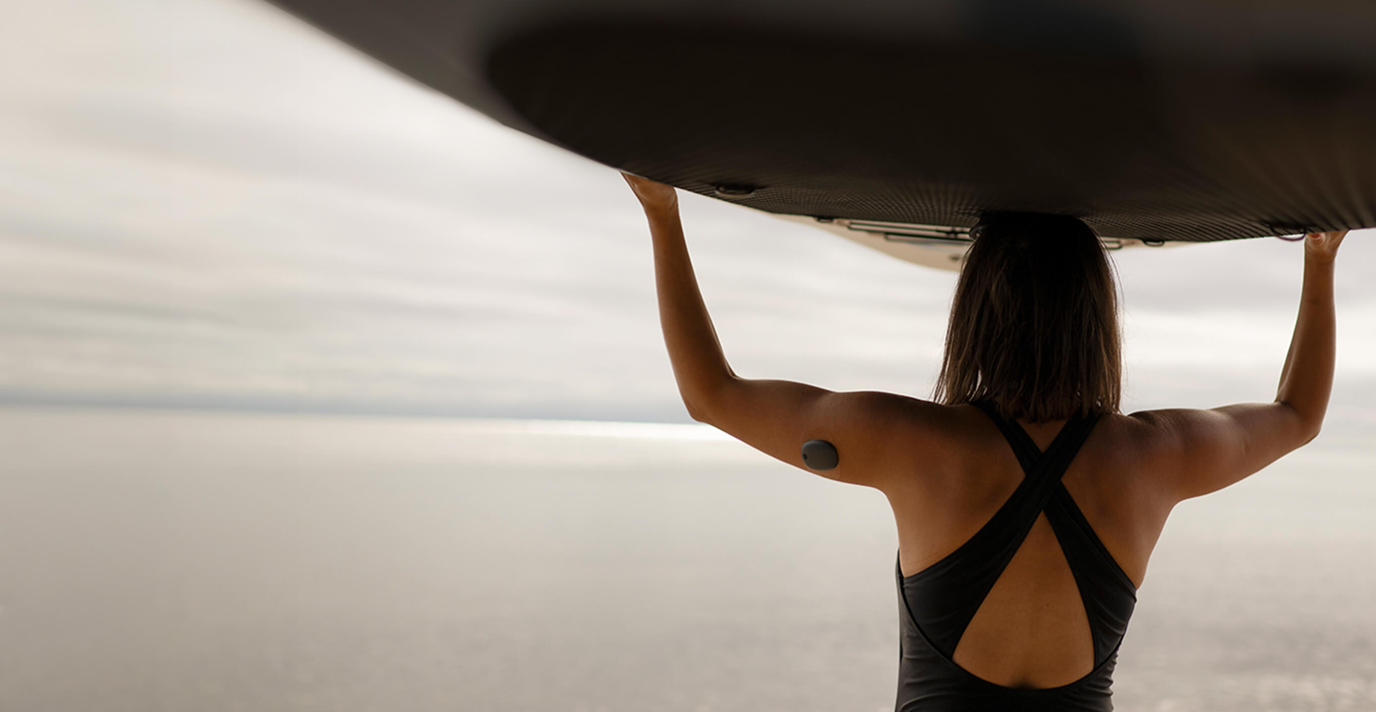 Kvinnlig surfare med Eversense-sändare på armen ser ut över havet medan hon bär sin surfbräda på huvudet