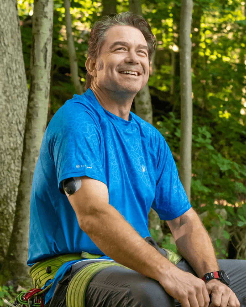 En man i en blå skjorta och en Eversense glukosmätare ler medan han sitter i en solig skog.