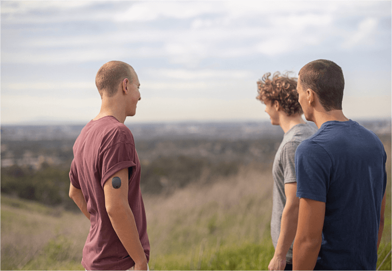 3 vänner står utomhus i ett fält och en av dem bär Eversense CGM-sändare
