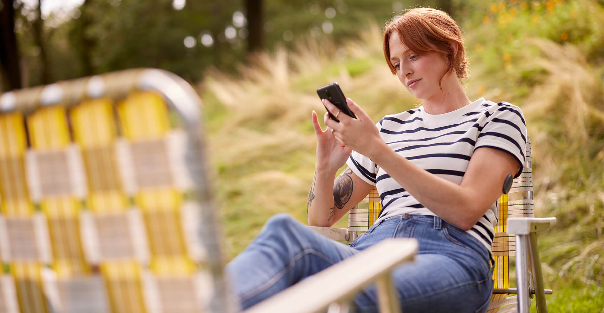 En ung kvinna sitter i en trädgårdsstol ute i en blommig trädgård och ser på sin mobiltelefon med en Eversense-sändare på armen