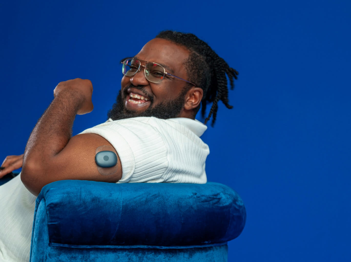 Eversense-användare som ler och böjer en arm med en CGM-sensor på armen, sittande på en blå stol.