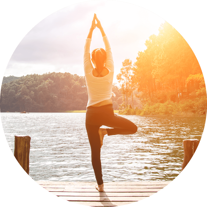 Eversense-användare som gör yoga-trädposition på en träbrygga, omgiven av en lugn sjö och skogsklädda kullar, med solljus.