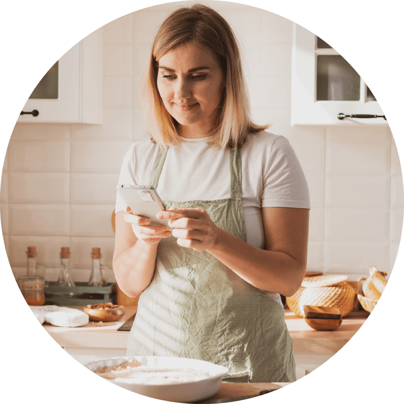 Eversense CGM-användare i ett förkläde använder en smartphone i ett ljust kök, med bakingredienser på bänken.